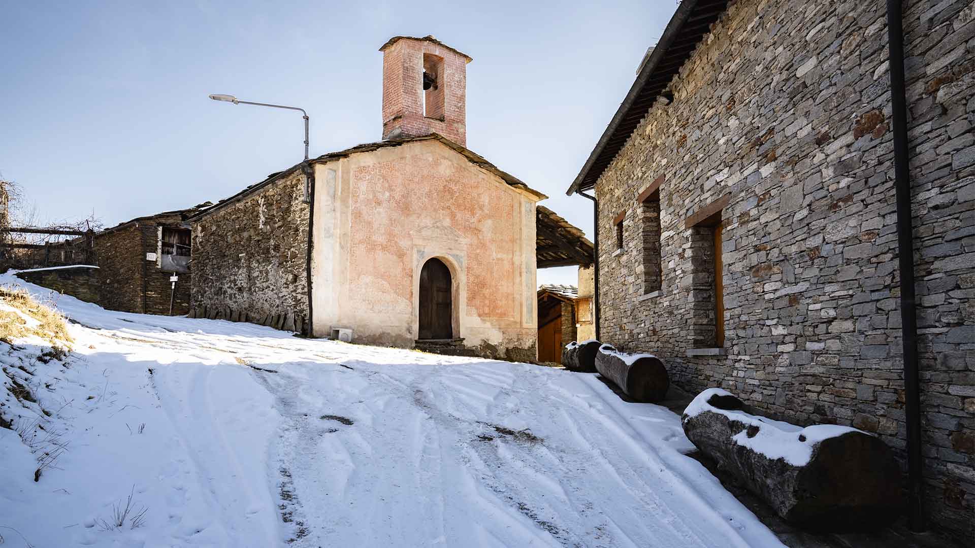 Cappella in pietra con campanile e portale in laterizio rosa innevata, edifici tradizionali adiacenti a Ostana